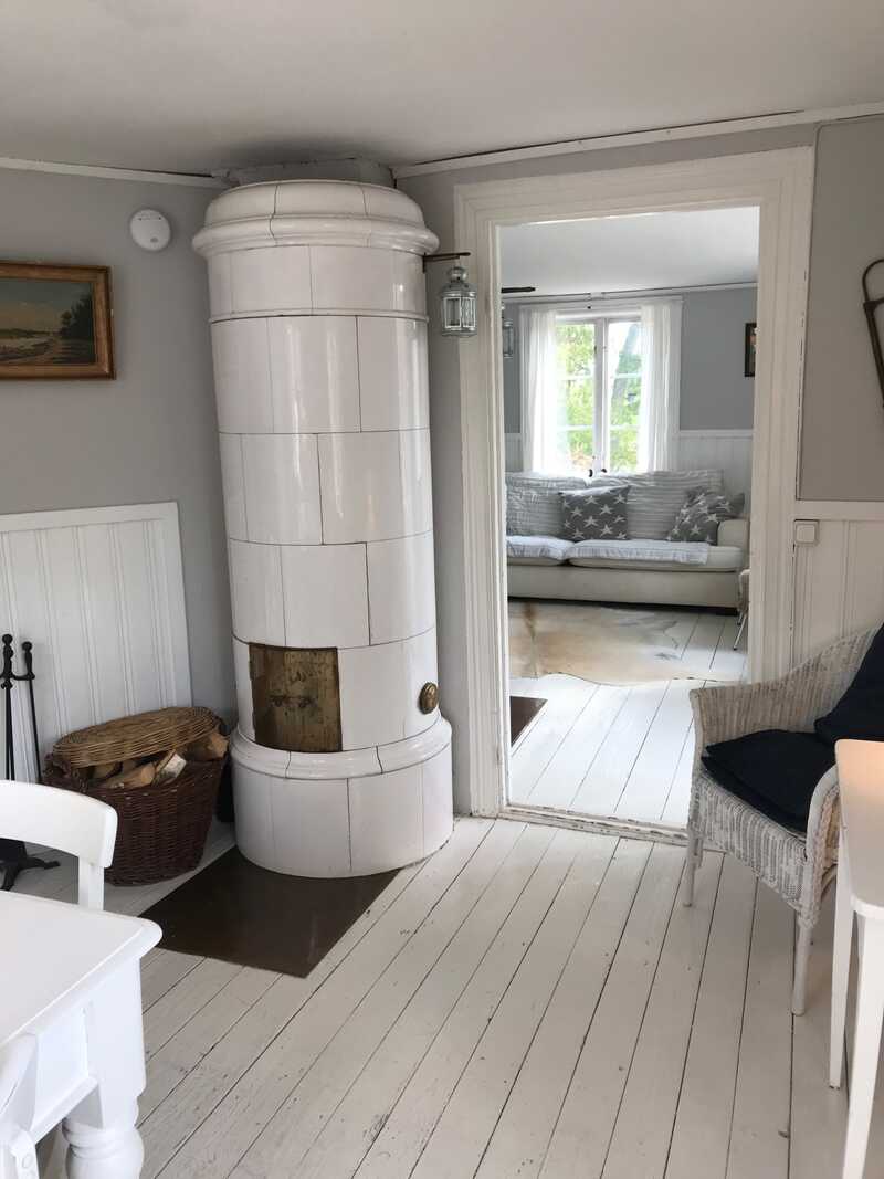 Round white tiled stove in a corner, with a basket of firewood beside it and a doorway leading to a sitting room